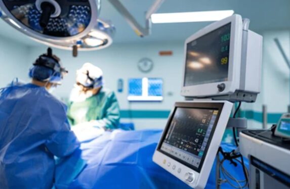 In an operating room, surgeons in blue scrubs perform surgery under bright overhead lights, with anesthesia and patient monitors visible in the foreground.